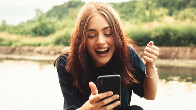 Happy bright smiling young woman sitting next to a river outdoors looking down to her phone face timing a friend