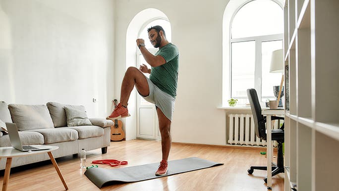 A man exercises on a yoga mat at home watching a pump exercise video. 