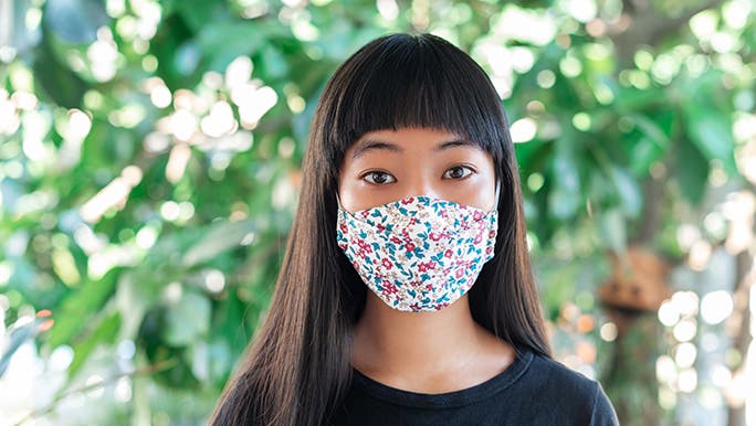 A woman looks straight on into the camera - she is wearing a mask and standing in front of a bunch of green plants. 