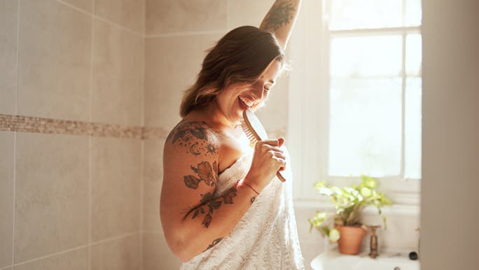 A happy-in-life woman sings into a wooden hairbrush in a sunny bathroom. She is wrapped in a towel and has a floral tattoo sleeve.
