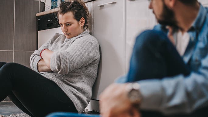 A woman and man sit on the floor of a kitchen, she has her arms crossed in front of her chest. It looks like there is a lot of conflict at the moment. 