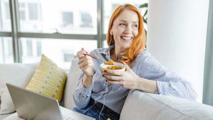 A young woman with red hair is sitting on a sofa with a laptop and smiling as she eats a bowl of corn flakes