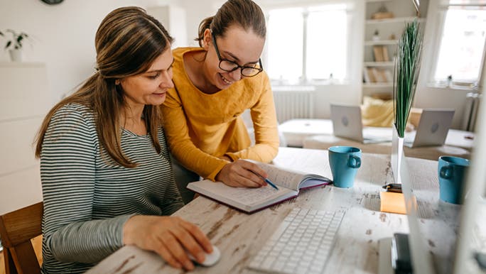 A woman and a teenage girl are in front of a computer together, they are writing in a life planner notebook and updating their work and life planners. 