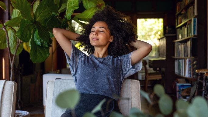 A young black woman is sitting on a sofa with her arms behind her head and her eyes closed