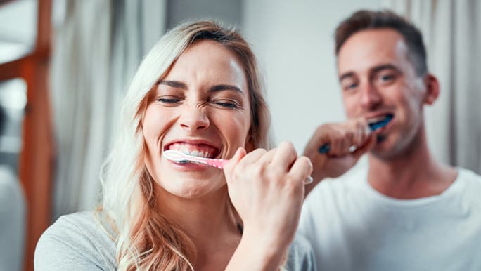 A couple smile and laugh while brushing their teeth as they debate whether it's better to brush their teeth before or after breakfast. 