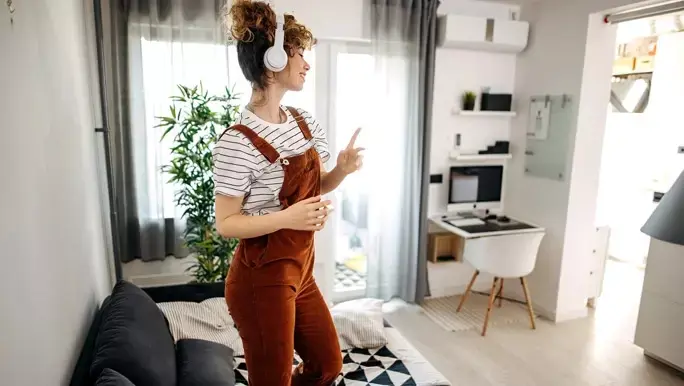Woman with curly hair wearing overalls and a striped tee dancing in the living room