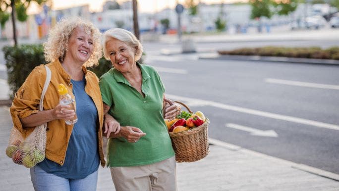 Two senior women are walking outdoors on a city footpath smiling as they link arms and hold shopping baskets and bags