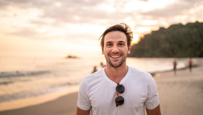A young man wearing a white T-shirt is standing on a beach and smiling 