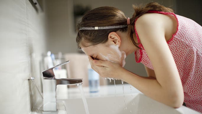 Teenage girl is washing her face and leaning over the sink. She’s removing the cleanser that’s on her face. 