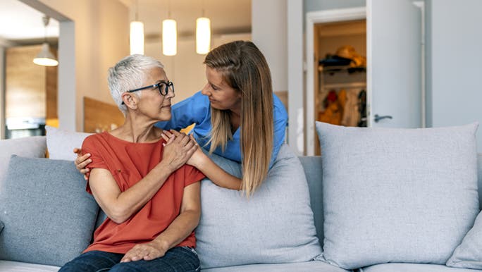 2 ladies who are friends, with one sitting on the couch, and the other standing behind her, reassuring her there’s no need to be scared of going to the doctor.