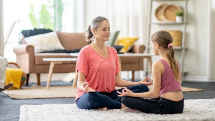A young mother and her daughter are sitting cross-legged on the floor and practicing mindfulness meditation together