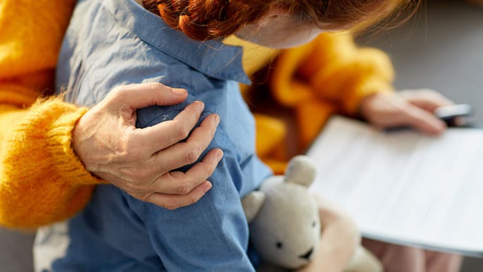 Up close, an adult arm is wrapped around a little girl’s shoulder. It looks like she is being comforted. The adult has some paperwork on her lap. 