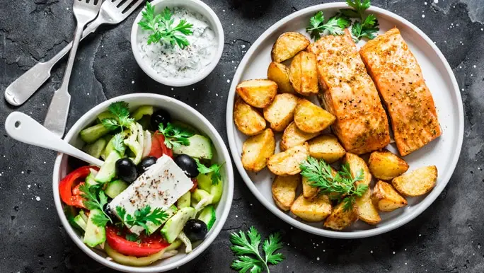 A bowl of Greek salad and a platter of roast potatoes and grilled salmon
