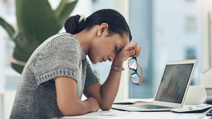 A woman with dark hair wearing a grey top and working in front of a laptop is leaning forward with her head in her hand, looking mentally exhausted