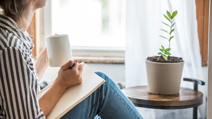 A woman writes in a notebook in a sunny corner of her home. She is wearing a striped shirt and drinking a cup of tea. 