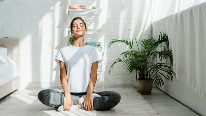 relaxed and happy woman with closed eyes practicing yoga in lotus position in bedroom in the morning