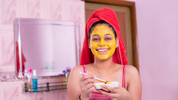 A young girl is applying a turmeric face mask while wearing a towel on her head and smiling