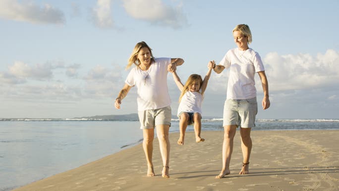 Two mums are holding their daughter’s hands and swinging her. They are happy on the beach.