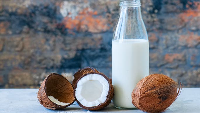 A jug of vegan coconut milk sits on a bench with some whole and halved coconuts.
