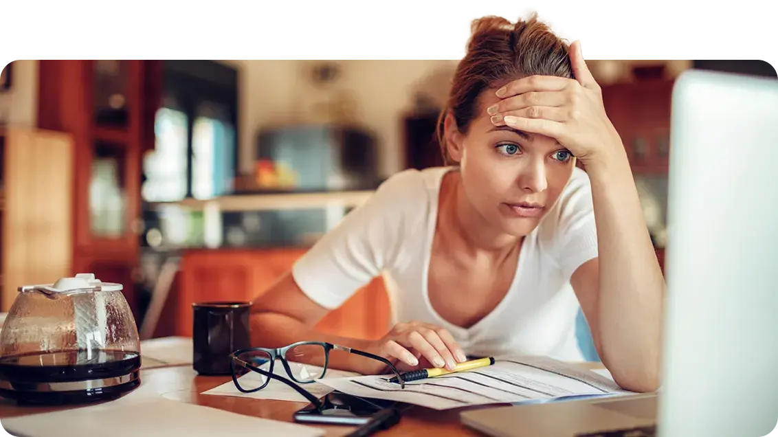 A woman sits in a cafe with a coffee pot, coffee mug, glasses, documents, pen and laptop at her desk. She is leaned forward looking at her laptop with the palm of her hand resting across her forehead. She looks stressed.
