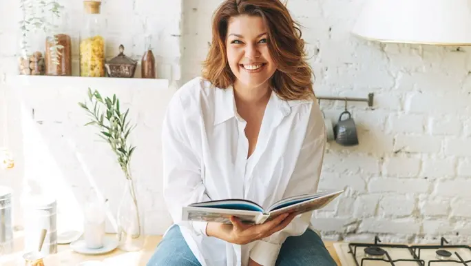 Happy young woman in blue jeans and white shirt reading cooking book in the home kitchen