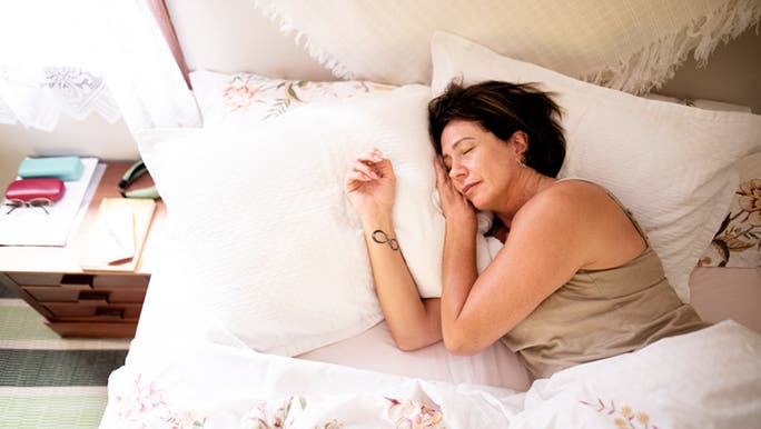 A woman with dark hair and a tattoo on her wrist is sleeping in a bed with light pouring in through the window
