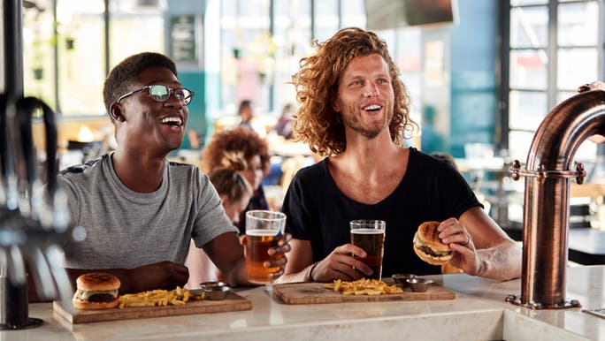  A dark-skinned man and a Caucasian man are smiling and eating burgers and chips and drinking beer