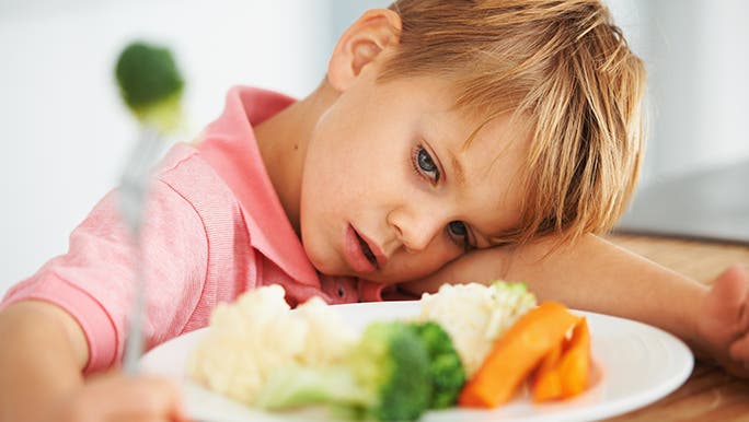 Little boy with his head on the table next to a plate full of vegetables. He is a picky eater who doesn’t like vegetables. 