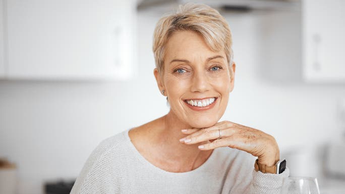 A middle-aged woman with short blonde hair is holding her chin in her hand and smiling at the camera