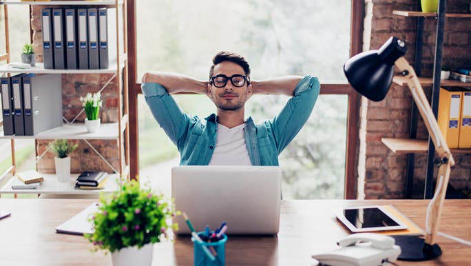 A man wearing black glasses and a blue shirt is sitting in front of a laptop with his arms behinds his head and his eyes closed