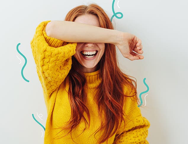 Young woman with long red hair in a yellow sweater covering her eyes with her arm and laughing