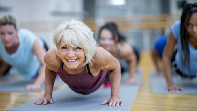A woman is doing a plank pose on a Yoga mat in an exercise class. She looks very healthy for her age. 