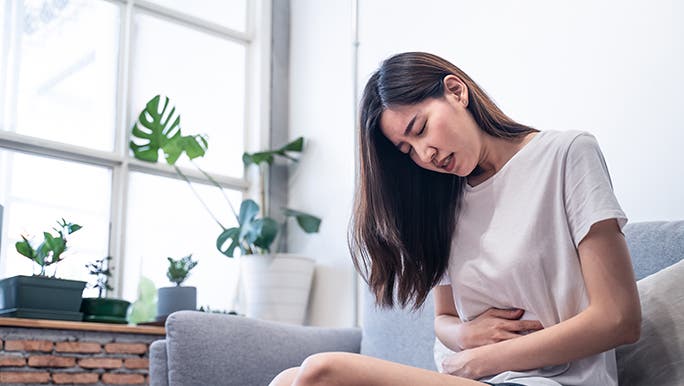 A woman sits on her couch and holds her stomach. She may have been eating foods that don’t promote gut health. 