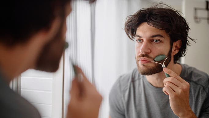 A young Caucasian man is looking at himself in a mirror and using a jade face roller to massage the skin on his face