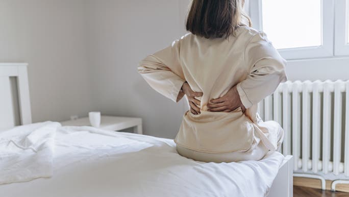 A woman in silk pyjamas is sitting on the side of a bed, holding her lower back because her muscles are sore. 