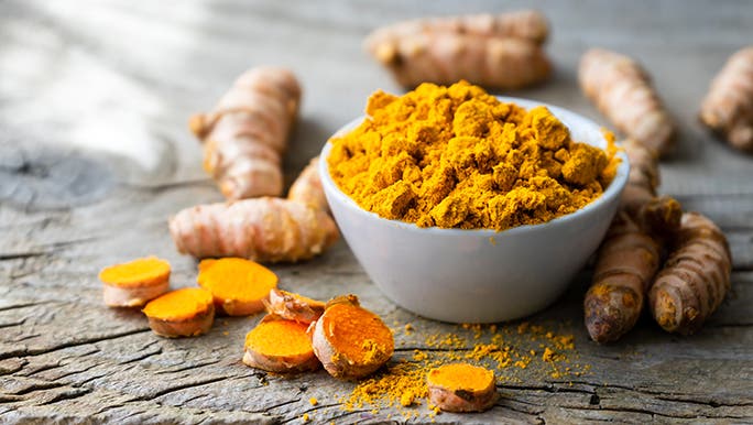 A bowl of ground turmeric piled in a white bowl with whole pieces of raw turmeric on a wooden table.