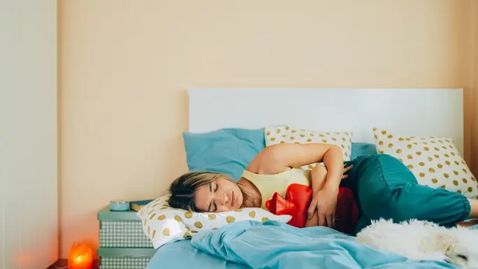Woman with abdominal cramps lying on her bed with a red hot water bottle