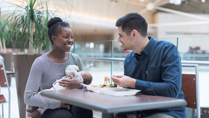 A woman sitting at a table with her partner in a food court, breastfeeding her child and discussing breastfeeding foods.