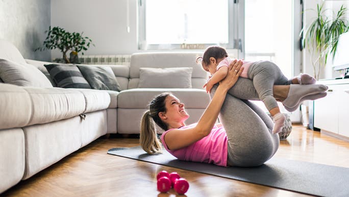 A mother exercises at home with her baby, she is doing crunches with her baby on her knees. 