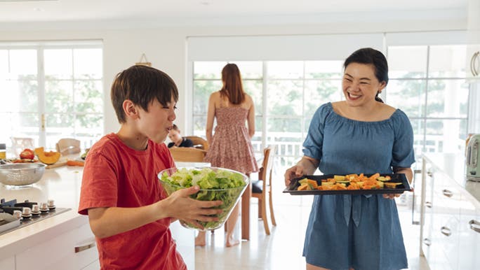 A mother and her son are in the kitchen standing as they prepare to walk to the dining table, his mother with a tray of healthy food in her hands. And her son, holding a bowl of salad.  