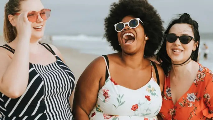 Three friends on a beach smile and laugh together to celebrate Galentine’s Day