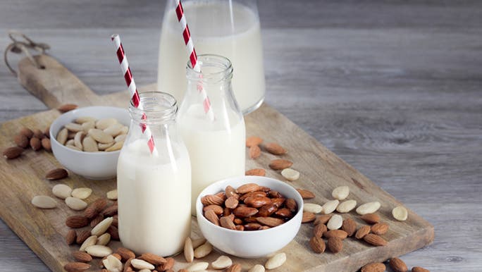 Two glasses of almond milk sit in front of a jug on a wooden chopping board with almonds sprinkled around which may be someone’s best milk alternative. 