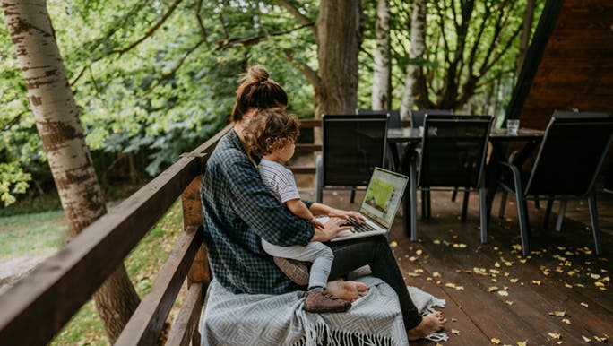 A mother works on a shady deck with a child on her lap. 