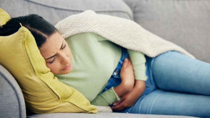 A young woman is lying on a sofa clutching her tummy as if she has gut pain