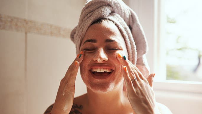 A woman with tattoos and bright fingernails is in a bathroom applying glycolic acid for sensitive skin.