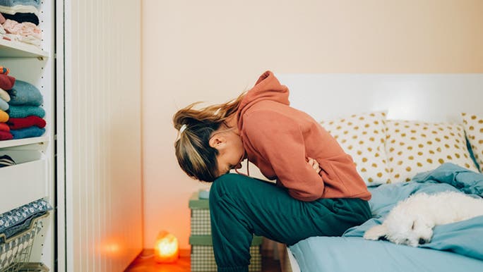 Young woman wearing an orange hoodie is doubled over on her bed clutching her stomach as if in pain from IBS, which may be helped by following a diet on the low FODMAP food list