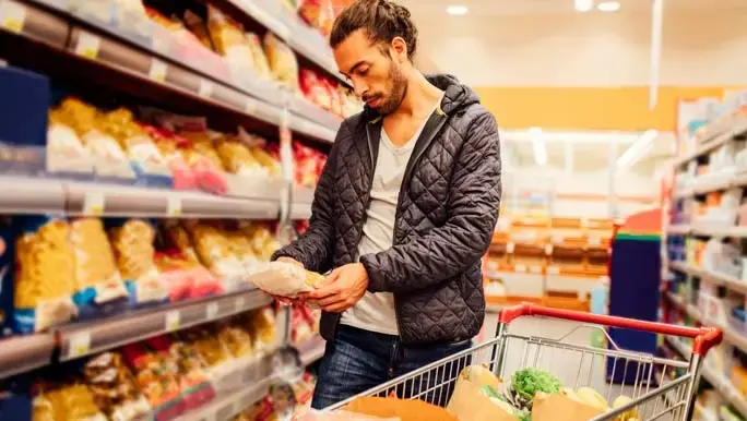 Young bearded man doing grocery shopping. He is choosing pasta and reading the nutrition label on the product