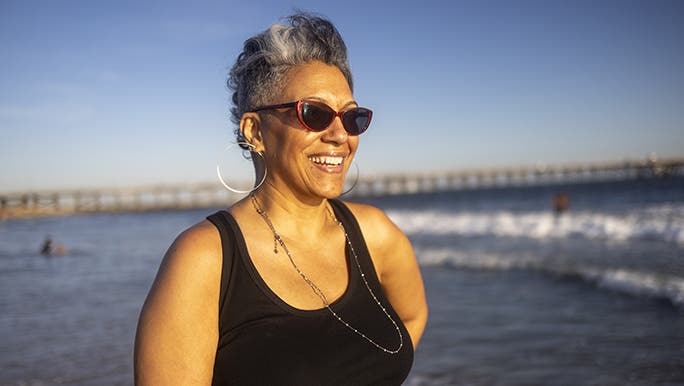A woman is smiling at the beach, she wears a black singlet and red sunglasses.