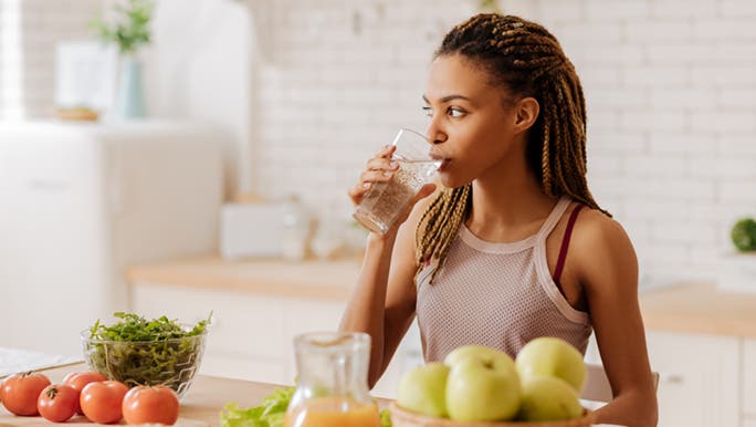 A lady is standing in front of her kitchen bench, drinking a tall glass of water, before she eats her breakfast. Perhaps more water and a healthy diet is one of the ways to look good without makeup.