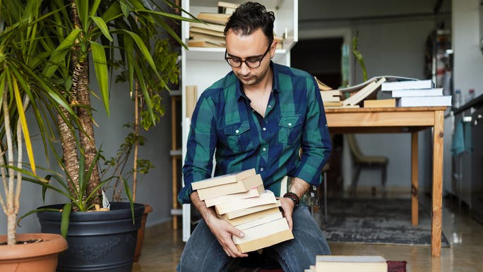A young man in a green checked shirt is sitting on the floor tidying a stack of books